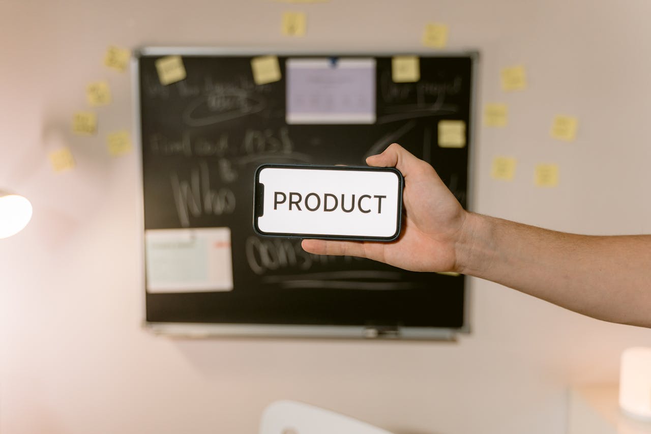 A hand holds a smartphone displaying the word PRODUCT in front of a blurred office board.