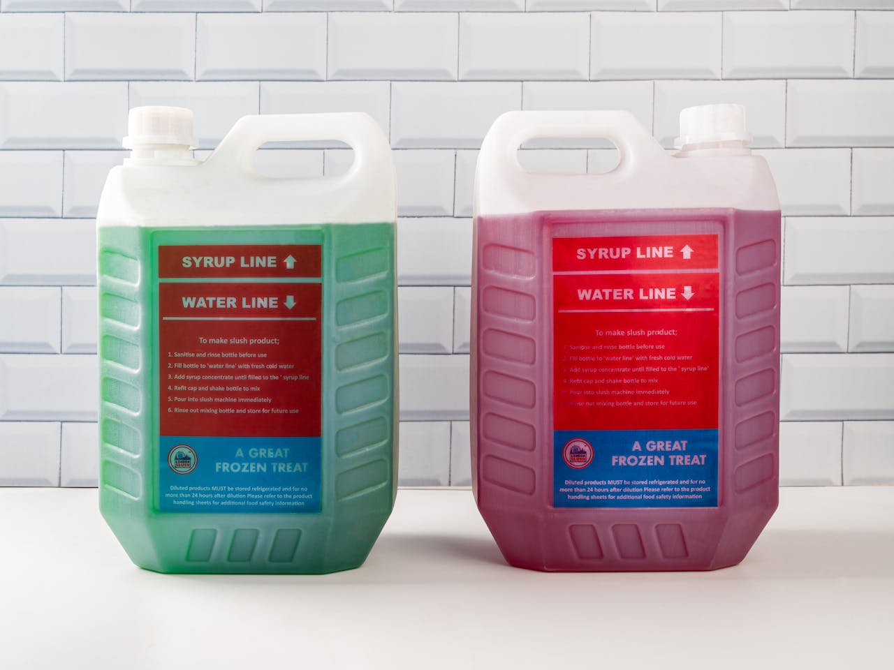 Two vibrant slush syrup containers with distinct colors and clear labels against a tiled wall backdrop.