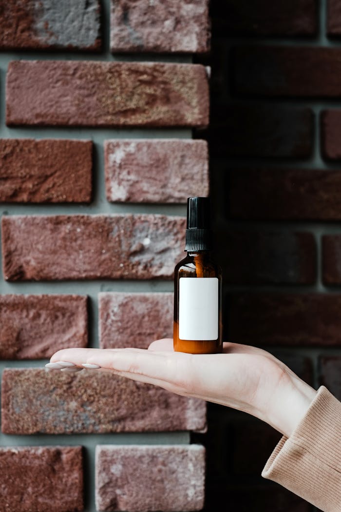 Hand holding a beauty product bottle with a blank label against a rustic brick wall backdrop.